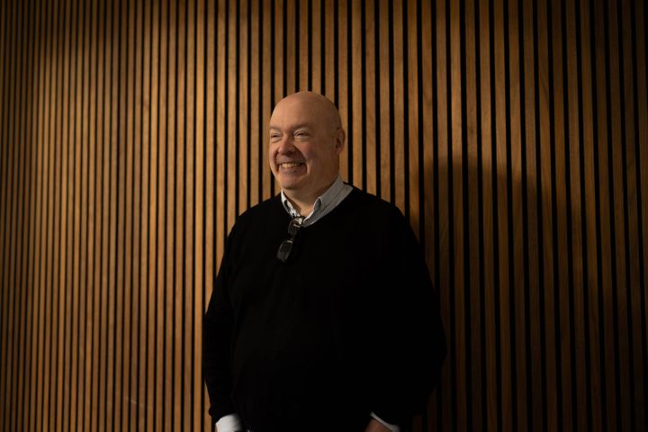 Phil stands in front of the wooden walls of a lecture theatre, smiling, in a black jumper.