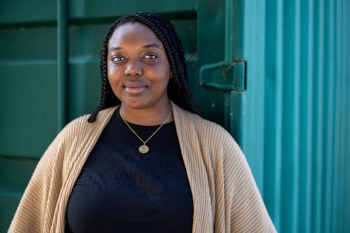 Evo stands against a green shipping container. She wears a beige cardigan and a gold necklace, and smiles at the camera.