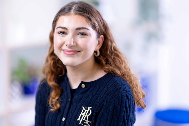 Irena wears a blue cardigan and gold earrings. She sits, smiling at the camera.