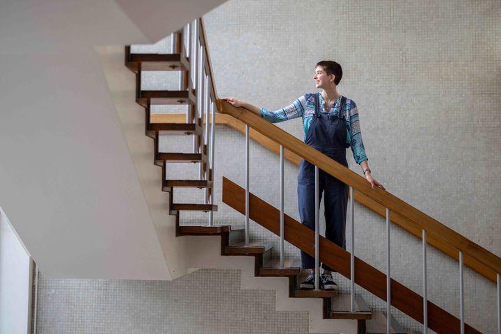 Vittoria stands with her hands on the railing in a stairwell, looking out towards the window.